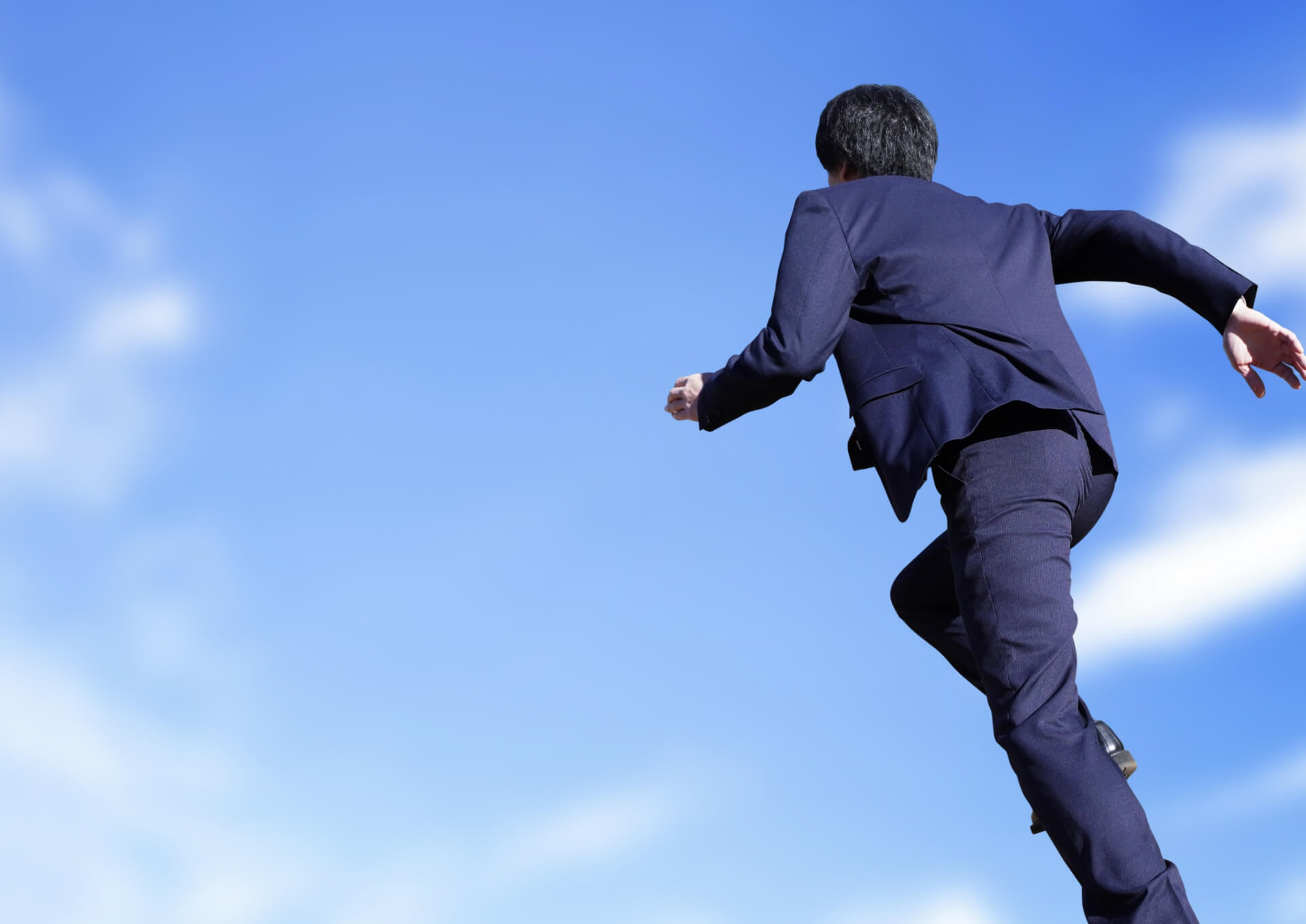 青空に向かって力強く跳躍するビジネスマンの後ろ姿。企業の成長を自らの力で推進する、自走する社員のイメージ。